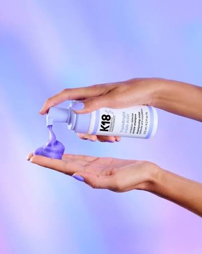 A person's hands holding a bottle of K18 hair color and dispensing purple hair dye onto a hand, set against a soft purple and blue gradient background.